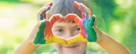 Ett barn med färgglada målade händer formar ett hjärta framför sitt ansikte utomhus.