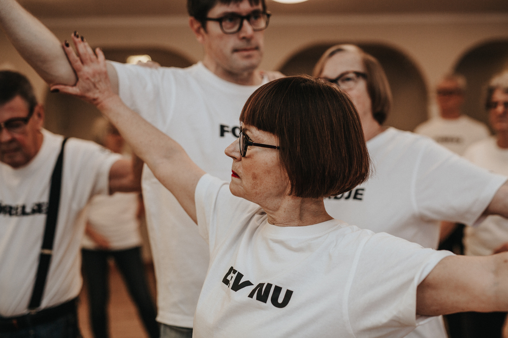En grupp äldre vuxna i vita t-shirts deltar i en dans- eller rörelseklass inomhus.