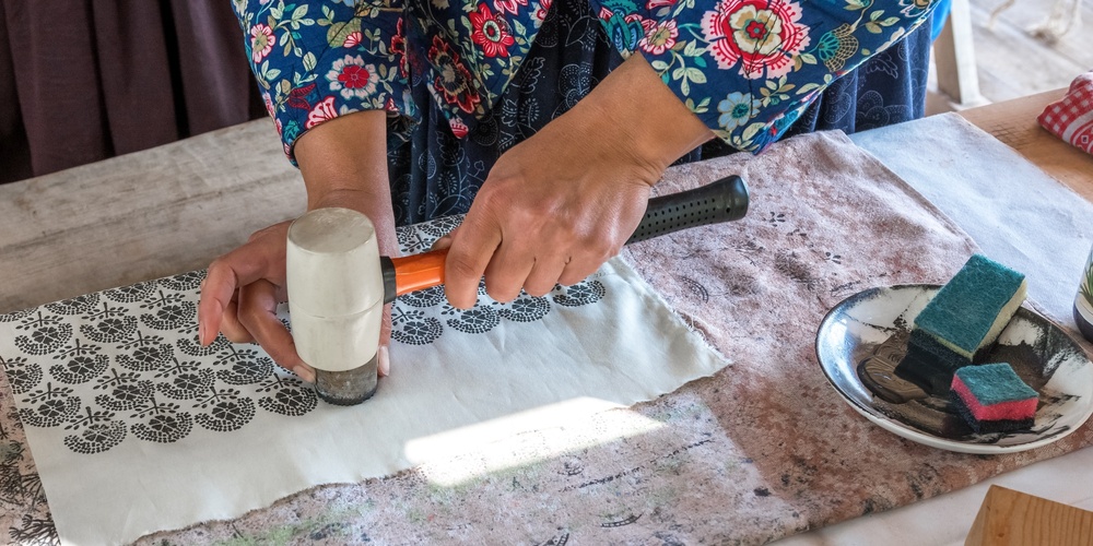 En person trycker mönster på tyg med hjälp av en stämpel och hammare vid ett bord.