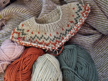 En stickad tröja i naturfärger under arbete, omgiven av garnnystan i grönt, orange, beige och rosa.