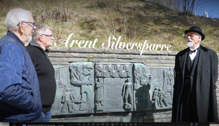 Tre män samtalar vid en relief med texten 'Arent Silversparre' utomhus på en solig dag.