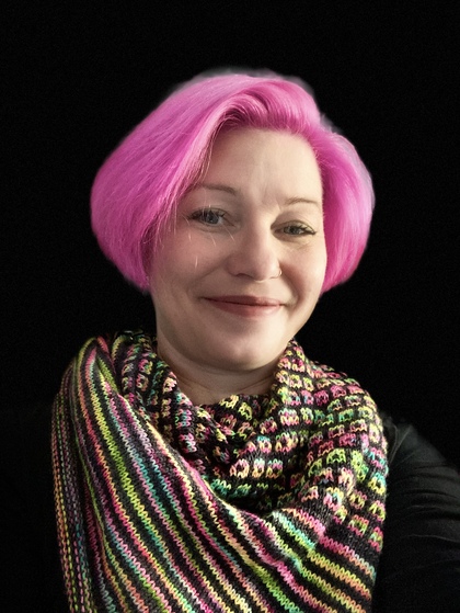 Leende person med kort, rosa hår och färgglad stickad sjal mot svart bakgrund.