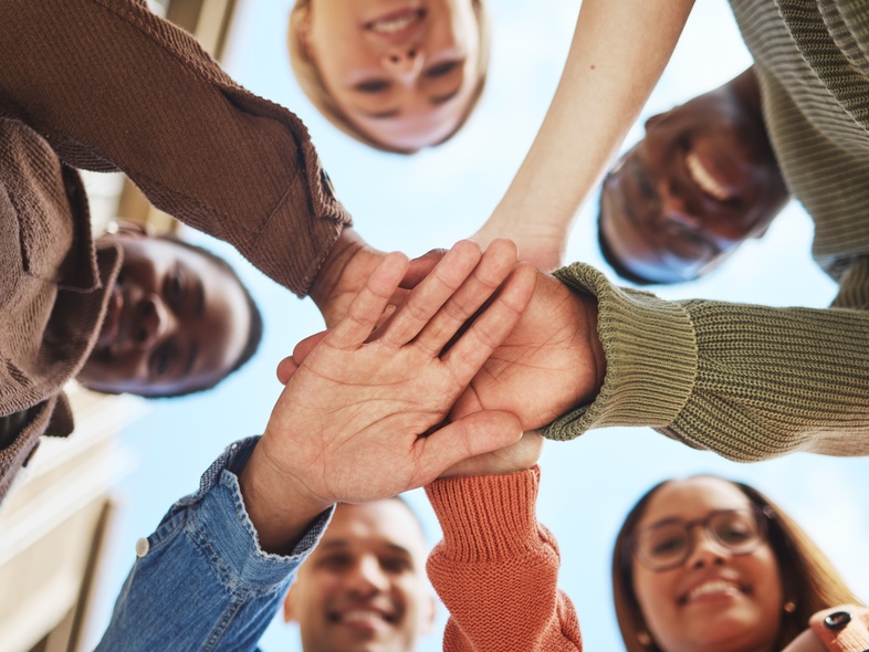 Fem personer står i en cirkel och lägger sina händer ovanpå varandra, sedda underifrån mot himlen.
