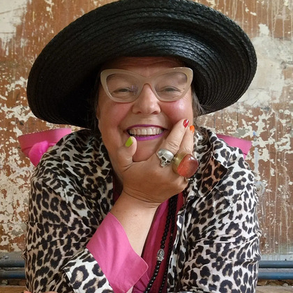 Leende person med stor svart hatt, glasögon och leopardmönstrad jacka mot sliten vägg.