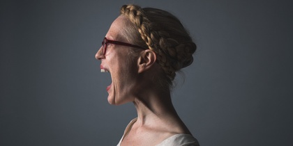 Kvinna med flätat hår och glasögon skriker i profil mot en mörk bakgrund.