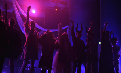 En grupp personer står på en scen med upplyst bakgrund i lila och rosa, med armarna höjda i luften.