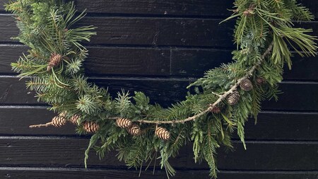 En halvcirkelformad grön krans med kottar hänger mot en svart trävägg.