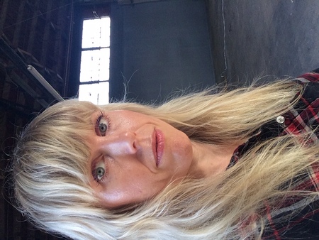 Kvinna med långt blont hår och lugg står i en tom industrilokal med stora fönster i bakgrunden.