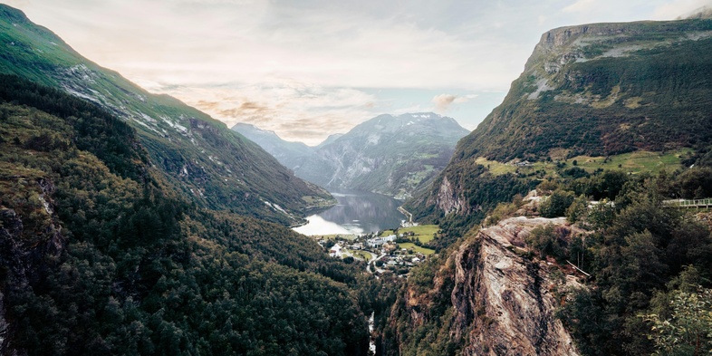 Bild från Norge, utsikt över en djup dal med gröna berg, en by och en sjö i fjärran under en molnig himmel.