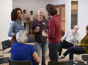 En grupp vuxna samtalar och dricker kaffe i en ljus samlingslokal med stolar i en cirkel.