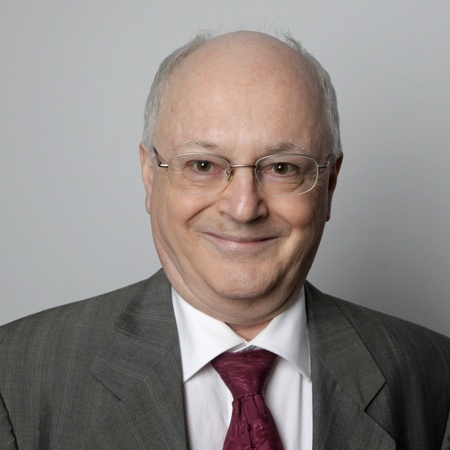 Äldre man med glasögon, grå kostym och röd slips ler mot kameran framför en ljusgrå bakgrund.