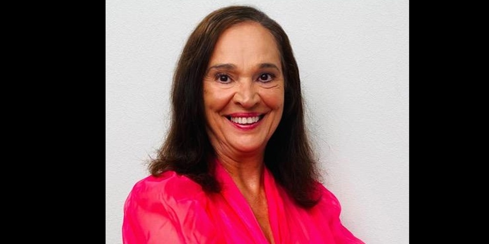 Person med långt mörkt hår i en starkt rosa blus står med armarna i kors mot en vit bakgrund.