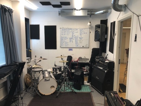 Ett musikrum med trumset, förstärkare, högtalare och en whiteboard på väggen.