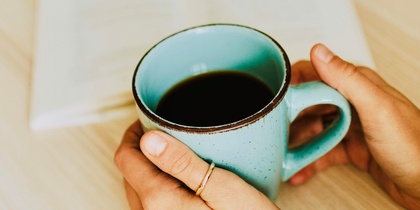 En person håller en blå mugg med kaffe framför en öppen bok på ett bord med pumpor i bakgrunden.