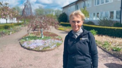 Person står i en trädgård med blomsterbäddar och gångvägar, byggnad i bakgrunden.