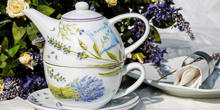 En tekanna och kopp med lavendelmotiv står på ett fat, omgiven av blommor och en servett på tallrik.