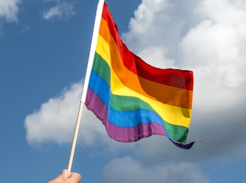 En hand håller upp en regnbågsflagga mot en blå himmel med vita moln.