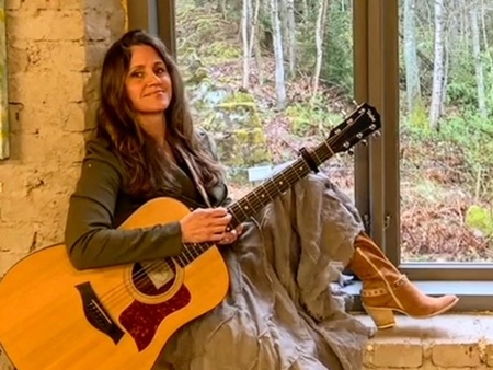 Kvinna sitter på en fönsterbräda med en akustisk gitarr, klädd i lång klänning och boots, inomhus vid tegelvägg.