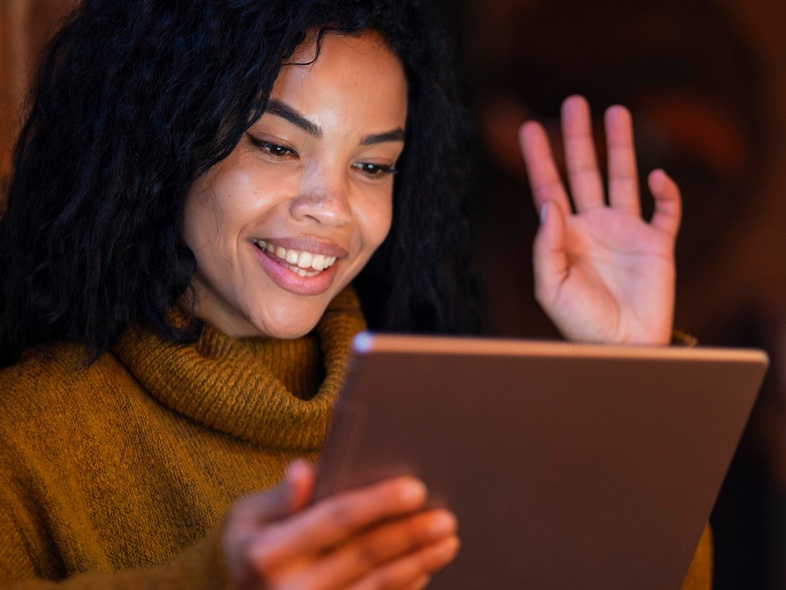 Person med mörkt lockigt hår och brun polotröja vinkar och ler mot en surfplatta i ett varmt upplyst rum.