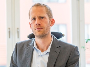 Man i kostym sitter på ett kontor framför ett fönster, tittar mot kameran.