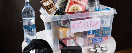 En plastlåda märkt 'KRISLÅDA' fylld med mat, vattenflaskor och nödvändiga saker står på ett runt bord.