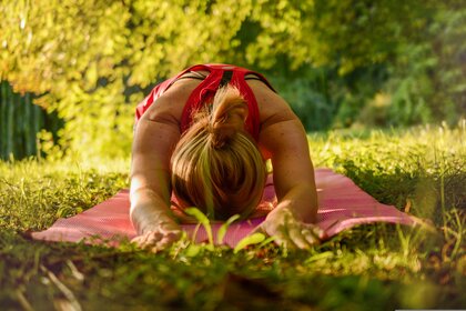 Person gör yoga i barnets position på en rosa matta utomhus i grönskande miljö.