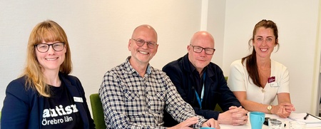 Fyra personer sitter vid ett konferensbord med anteckningsblock, broschyrer och kaffekoppar.