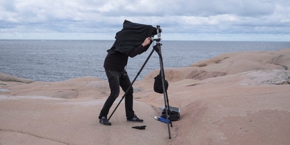 En person fotograferar med en stor kamera på stativ vid havet, täckt av en svart duk.