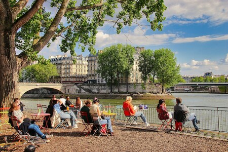 Folk sitter på uteservering vid floden med utsikt över bro, träd och klassiska byggnader i solsken.