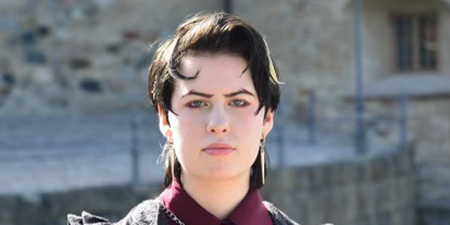 person med svart hår och röd krage som står framför grå tegelbyggnad