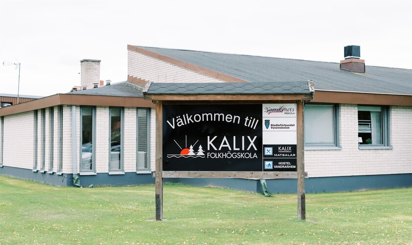 Skylt med texten 'Välkommen till Kalix folkhögskola' framför en låg, ljus byggnad med gräsmatta.