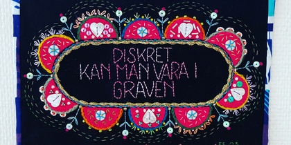 Broderad texttavla med färgglada mönster och texten 'Diskret kan man vara i graven' på svart bakgrund.