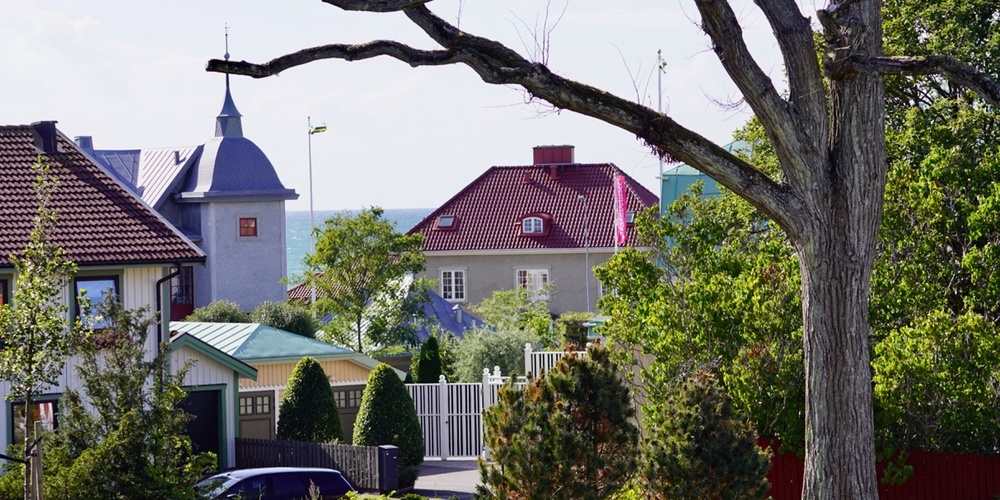Vy över ett bostadsområde med färgglada hus, trädgårdar och ett stort träd i förgrunden.