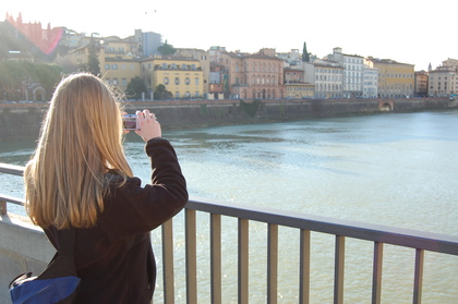 En person med långt blont hår fotograferar en stadsvy över en flod från en bro.