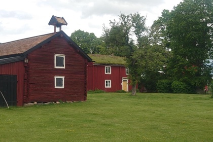 Två röda trähus på en grön gräsmatta med träd och molnig himmel i bakgrunden.