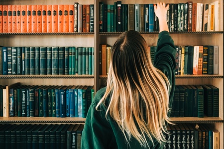 En person med långt blont hår sträcker sig efter en bok på en bokhylla fylld med böcker.