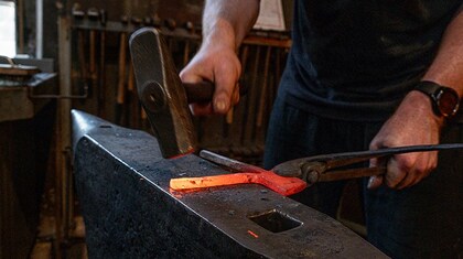 En person smider en glödande metallbit på ett städ med hammare och tång i en smedja.