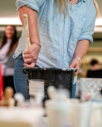 En person rör om i en svart hink med en träpinne, omgiven av olika burkar på ett bord.