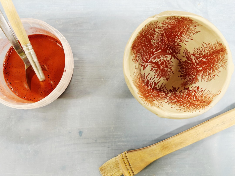 En skål med röd dekor, en pensel och en kopp med röd färg på ett bord.