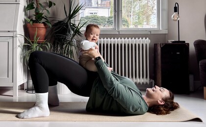 En person gör höftlyft på en yogamatta inomhus och håller ett litet barn på magen.