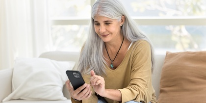 Kvinna med långt grått hår sitter i soffa och använder en smartphone.