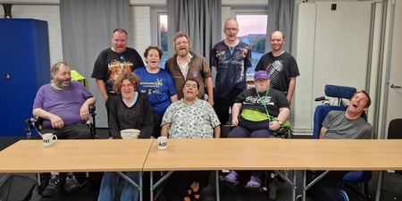 En grupp på tio personer, några i rullstol, sitter och står runt ett bord i ett mötesrum och skrattar tillsammans.