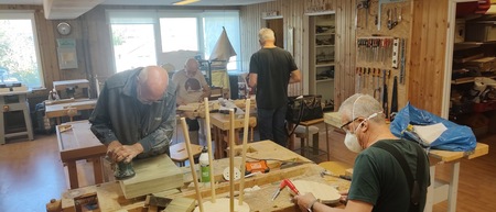 Fyra äldre män arbetar med träslöjd i en ljus snickarverkstad med träpaneler och verktyg på väggarna.