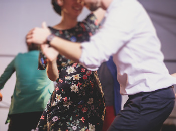 Två personer dansar tillsammans, en i blommig klänning och en i vit skjorta, i en suddig inomhusmiljö.