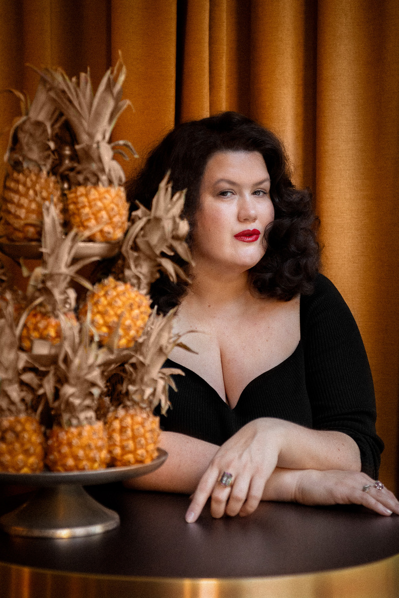 Maria Maunsbach med mörkt hår och röd läppstift sitter vid ett bord med en stapel av ananas framför sig, mot en guldfärgad gardin.