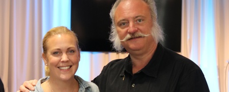 Två personer står tillsammans i ett rum med ljusa gardiner och en stor TV-skärm i bakgrunden.