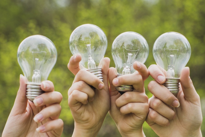 Fyra händer håller upp varsin glödlampa utomhus med grönskande bakgrund.