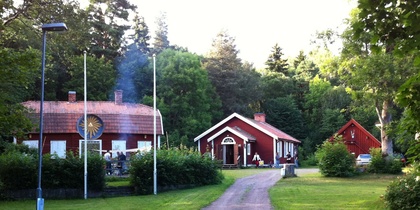 En grusväg leder till tre röda hus omgivna av grönska och träd, med flaggstänger och en grillplats.