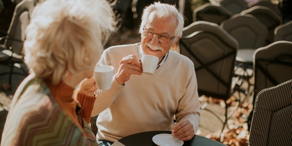 Två äldre personer sitter utomhus vid ett runt bord och dricker kaffe tillsammans.