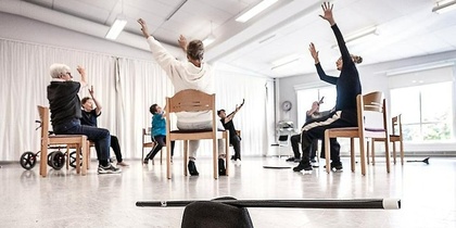 En grupp personer sitter på stolar i en ljus sal och sträcker armarna uppåt, hatt och käpp i förgrunden.